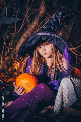 Witch girl in a hat with a pumpkin for Halloween. In the background there is a dense autumn forest.