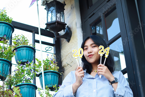 happy asian young woman hipster looking to camera with holding 2024 number candle celebrating new year eve, beautiful female standing in front of vintage house outdoor garden with low angle view photo