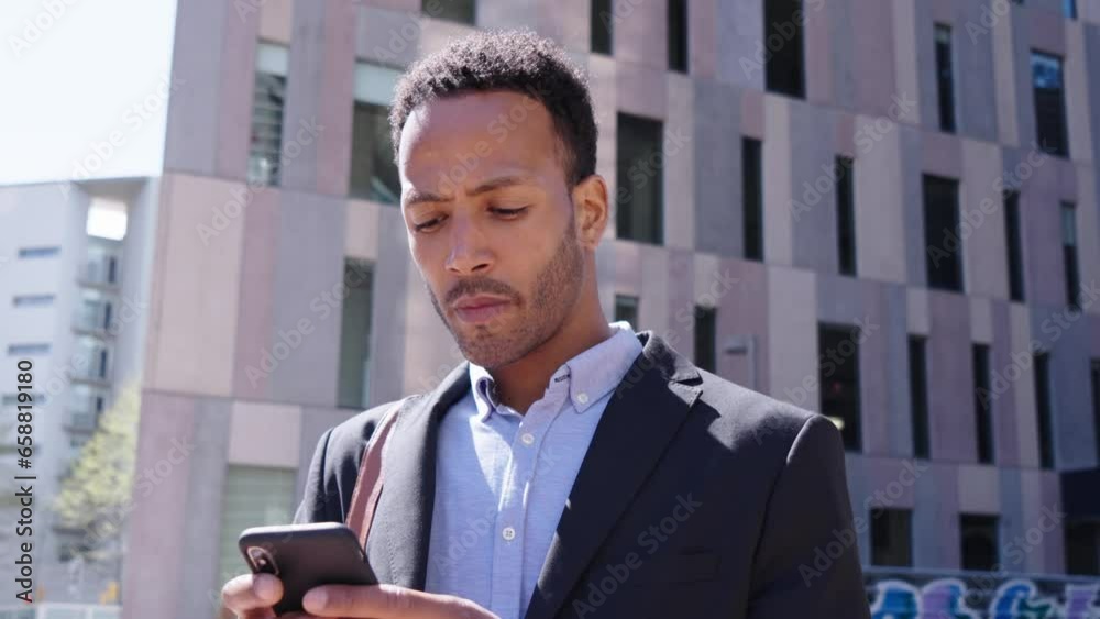 Serious young adult Latin professional male looks up pensive using smart phone center building background. Black business man in formal wear typing message online on mobile outside from office work.