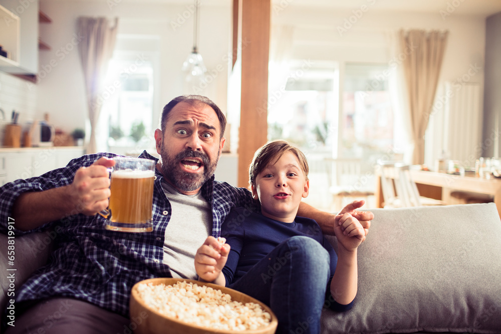 Young Caucasian father watching sports with his son and cheering for their team on the couch in the living room at home Young Caucasian father watching sports with his son and cheering for their team on the couch in the living room at home