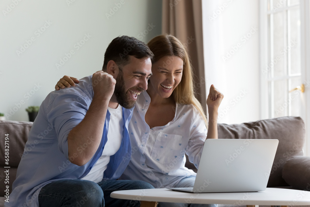 Excited overjoyed couple winning money prize, getting happy notice, making winner yes hands, laughing, screaming for joy, sitting on sofa, celebrating success at laptop at home - obrazy, fototapety, plakaty 