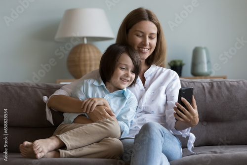 Happy beautiful mother and kid boy taking home selfie on smartphone, resting, hugging on couch, speaking on video call on mobile phone, enjoying online family communication