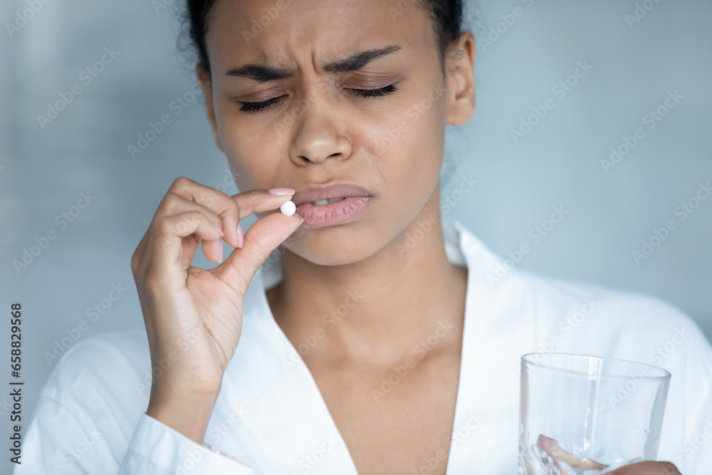 Unwell african American young woman feel sick suffer from migraine take antibiotic or aspirin pill with glass of water, unhealthy biracial female struggle with depression have antidepressant at home Unwell african American young woman feel sick suffer from migraine take antibiotic or aspirin pill with glass of water, unhealthy biracial female struggle with depression have antidepressant at home