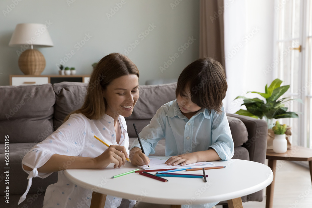 Cheerful pretty caring mother and cute little son boy drawing doodles in coloring pencils together, enjoying creative educational activity, family leisure time at home, smiling, talking Cheerful pretty caring mother and cute little son boy drawing doodles in coloring pencils together, enjoying creative educational activity, family leisure time at home, smiling, talking