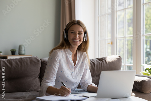 Happy cheerful millennial freelance business woman in headphones working at laptop from home, writing notes, talking to customer on video call, looking at camera, smiling, laughing, sitting on couch