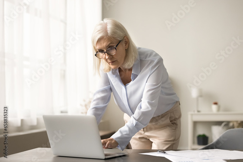Focused busy elderly business professional woman using laptop, standing at workplace table, typing on computer, thinking, reading on display, watching online content, working on business project
