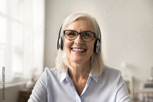 Happy pretty blonde older business woman in wireless headphones and glasses speaking on online conference, looking at camera with toothy smile, laughing. Video call screen head shot portrait