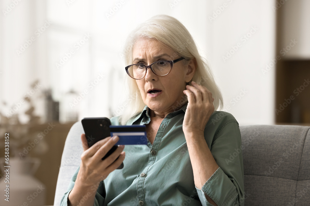 Worried surprised elderly woman in glasses looking at smartphone and credit card, getting financial problems, bad online bank service, finding overspending, bankruptcy risk Worried surprised elderly woman in glasses looking at smartphone and credit card, getting financial problems, bad online bank service, finding overspending, bankruptcy risk
