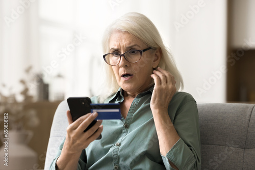 Worried surprised elderly woman in glasses looking at smartphone and credit card, getting financial problems, bad online bank service, finding overspending, bankruptcy risk