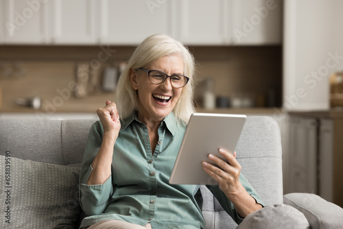 Cheerful excited elderly woman using digital tablet computer, making winner yes hand, laughing, getting great good news, looking at screen, enjoying success, triumph, win
