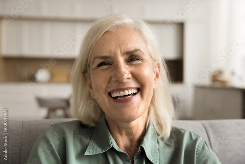 Joyful positive senior woman close up front head shot portrait. Happy blond old lady looking at camera with beautiful toothy smile, laughing, talking on video conference call at home
