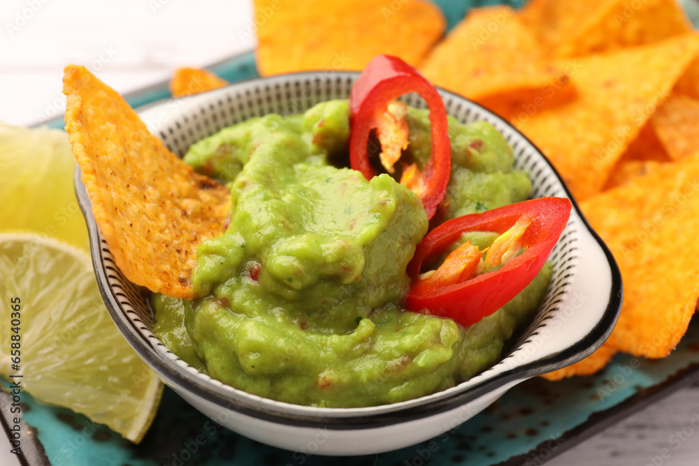 Bowl of delicious guacamole with chili pepper, nachos chips and lime on plate, closeup
