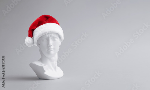 Trendy christmas composition made of fake statue of David's head with red Santa Claus hat on light background. Minimal concept of new year. Creative art, minimal aesthetics. With copy space for text.