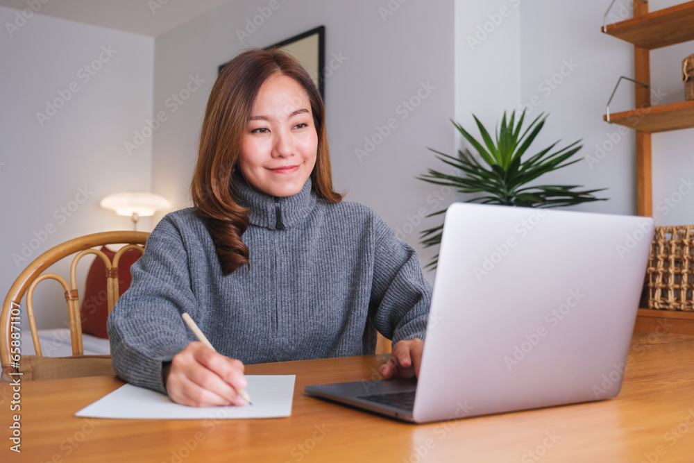 Portrait image of a young woman writing on paper while working on laptop computer at home