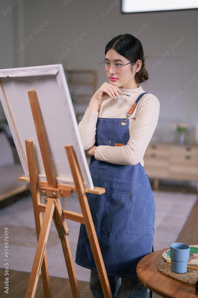 Young asian female artist hold paintbrush and looking on canvas to thinking create painting artwork