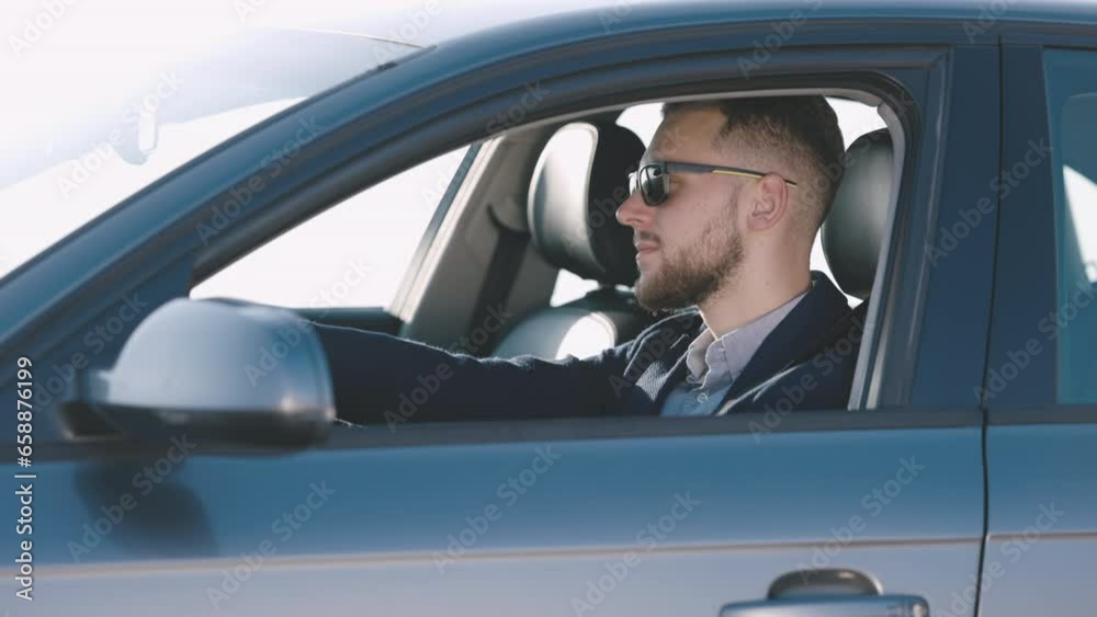Portrait of stylish young businessman man in glasses as drives car through bustling and sunlit city looking in mirror, view side Portrait of stylish young businessman man in glasses as drives car through bustling and sunlit city looking in mirror, view side