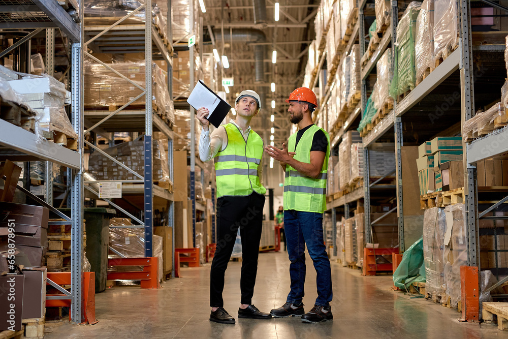 young Caucasian workers in uniform make an inventory management of products on shelves in warehouse, discuss together. Concept of good management system to support working with industrial business.