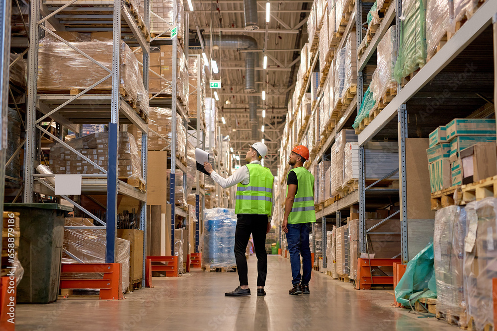 two men in warehouse wearing safety helmet, vest. Concept for industry, job, meeting, work training. Two caucasian warehouse workers walking in distribution storage area discussing two men in warehouse wearing safety helmet, vest. Concept for industry, job, meeting, work training. Two caucasian warehouse workers walking in distribution storage area discussing