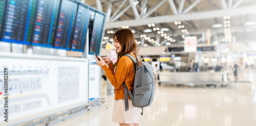 Young asian woman in international airport, using mobile smartphone and checking flight at the flight information board