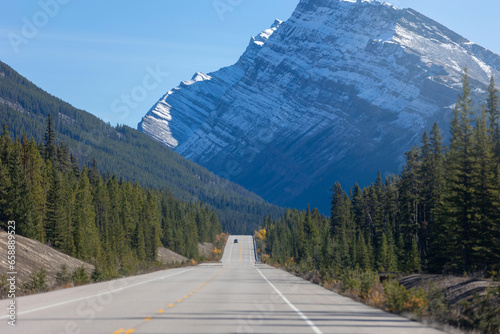 Beautiful scenery of a long straight road surrounded by high snowy rocky mountains, under blue sky