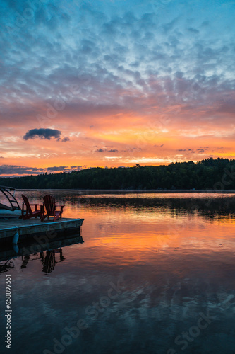 Two adirondack chairs on end of dock on lake at sunrise.