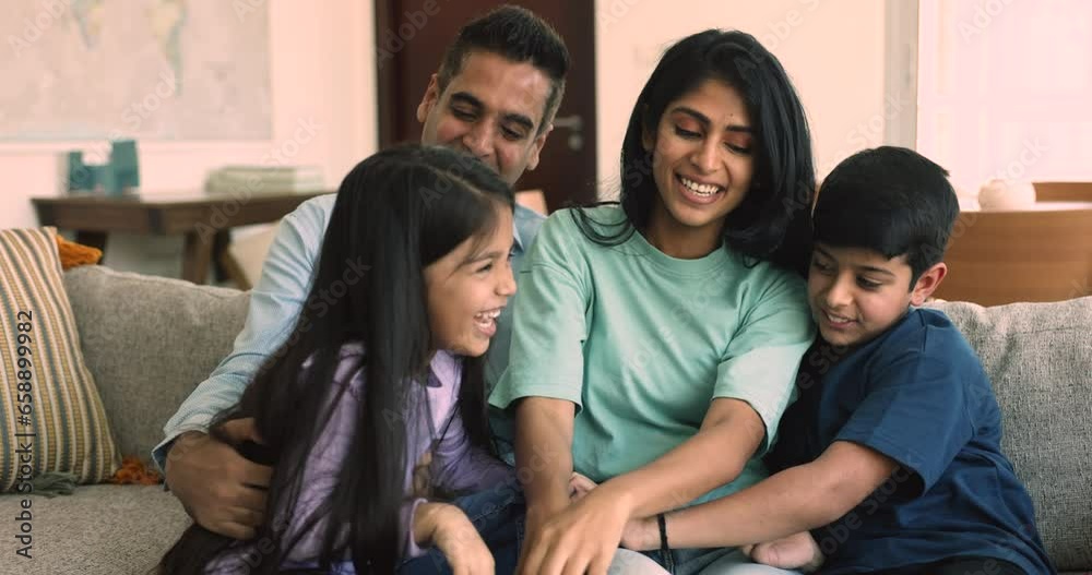 Beautiful Indian family with little children sit on sofa play together, tickling each other and laughing feel happy. Parents and kids spend time at home, enjoy playtime and warm relations. Ties, bond