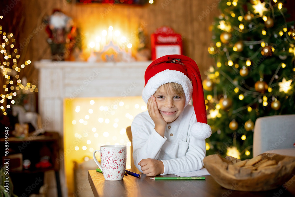Beautiful blond child, young school boy, writing letter to Santa Claus in a decorated home with knitted toys