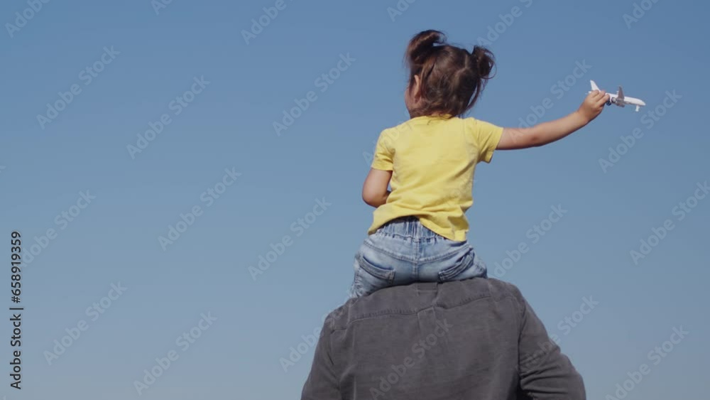 Little girl sitting on the shoulders of her father and playing with a toy plane, view from behind, while a real plane flies low over their heads in a clear blue sky Little girl sitting on the shoulders of her father and playing with a toy plane, view from behind, while a real plane flies low over their heads in a clear blue sky