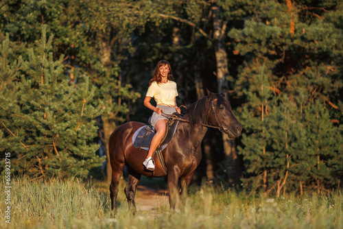 A young girl sits on a horse. Rider against the backdrop of the forest at sunset