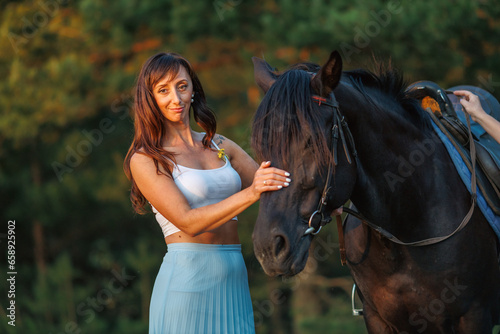 A young girl walks across a field. Portrait of a rider and a black horse against the background of a forest at sunset