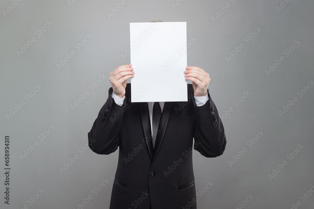 Business person wearing black suit hiding his face behind blank empty white paper on gray studio wall background Business person wearing black suit hiding his face behind blank empty white paper on gray studio wall background