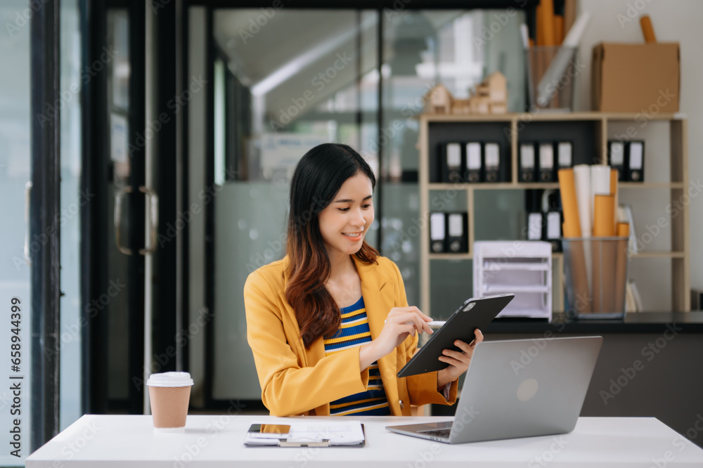 Confident business expert attractive smiling young woman typing laptop ang holding digital tablet on desk in office.
