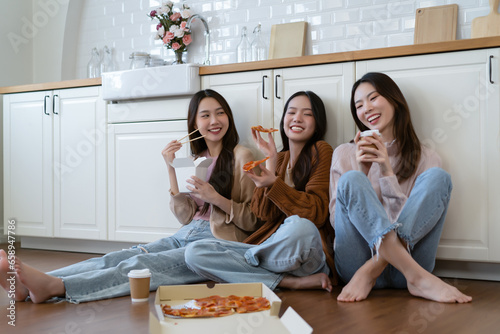Beautiful group of Asian friends wearing sweater sits on the floor enjoy eating pizza and takeaway food in kitchen room. Happy roommates having fun eating take away at home. Fast delivery food concept