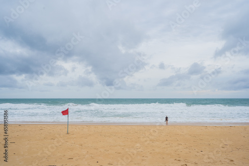 Female tourist standing on the beach  strong white waves and blue sky with white clouds  red flag symbolizing prohibition of swimming in the sea.