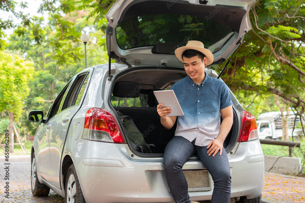 Asian man sitting back of car and holding tables check location and find place to travel. Happy road trip and vacation in summer in the countryside. Concept of journey on leisure, road trip vacation Asian man sitting back of car and holding tables check location and find place to travel. Happy road trip and vacation in summer in the countryside. Concept of journey on leisure, road trip vacation