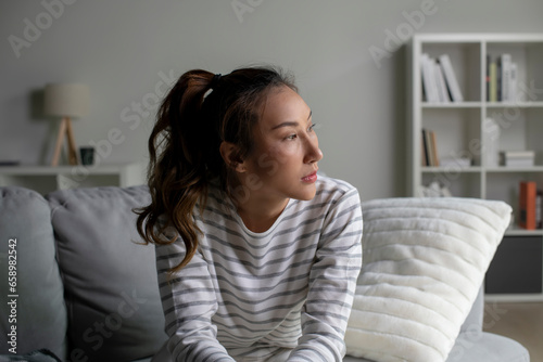 Lonely Asian woman in distress, worried, confused, sitting on sofa at home. Female suffer from mental health problems.