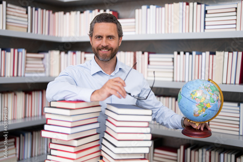 Portrait of teacher with book in library classroom. Handsome teacher in university library. Teachers Day. Teacher giving classes. School teacher in library. Tutor at college library on bookshelf.