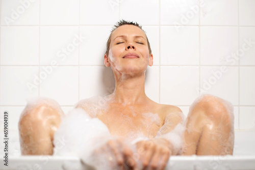 A middle-aged woman  about 40  smiles in a foamy bathtub. Her short hair is wet. Contentment fills her face. Pure bliss in the bath
