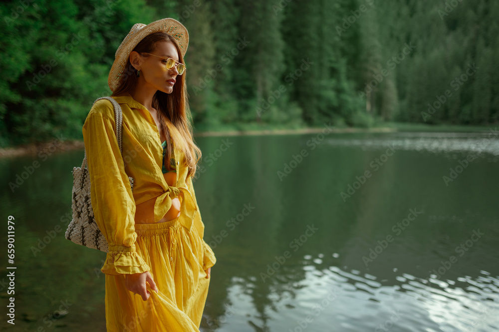 Fashionable woman wearing wicker hat, summer yellow suit with blouse and long skirt, sunglasses,  carrying stylish bag, posing near beautiful lake in mountains. Copy, empty space for text
