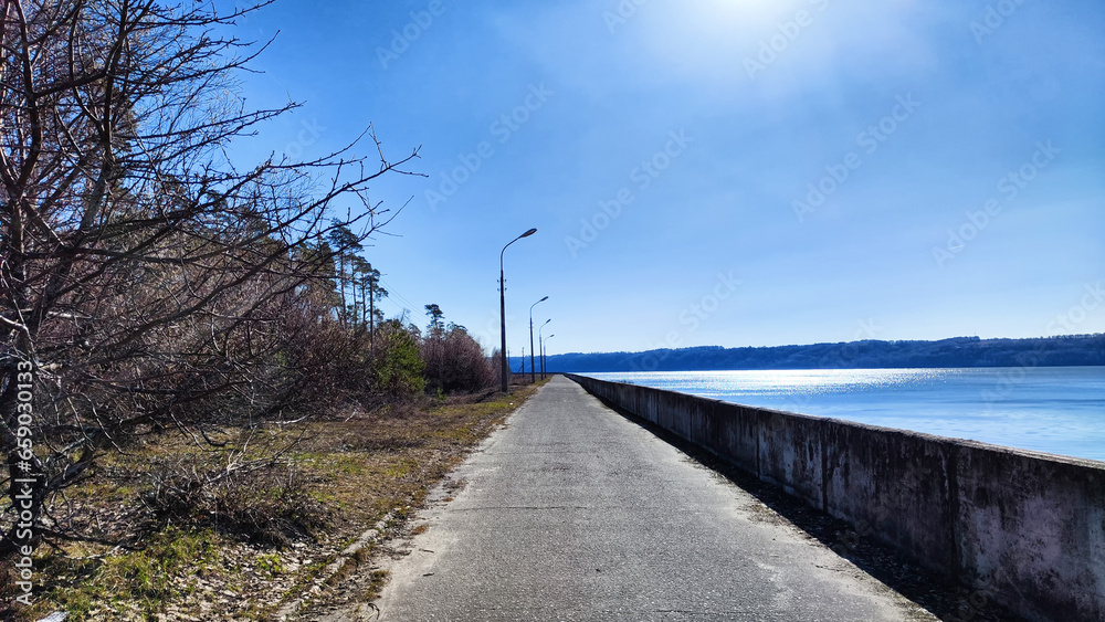 Asphalt path on the embankment near the water of the lake, sea, river, reservoir and blue distance with the sky with white clouds and horizon. Natural landscape with water on a sunny day