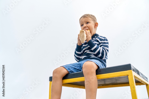 Hungry little boy with hood eats sandwich sitting on table on misty day in mountain park. Cute child bites snack resting in foggy highland. Kid has dinner outdoors