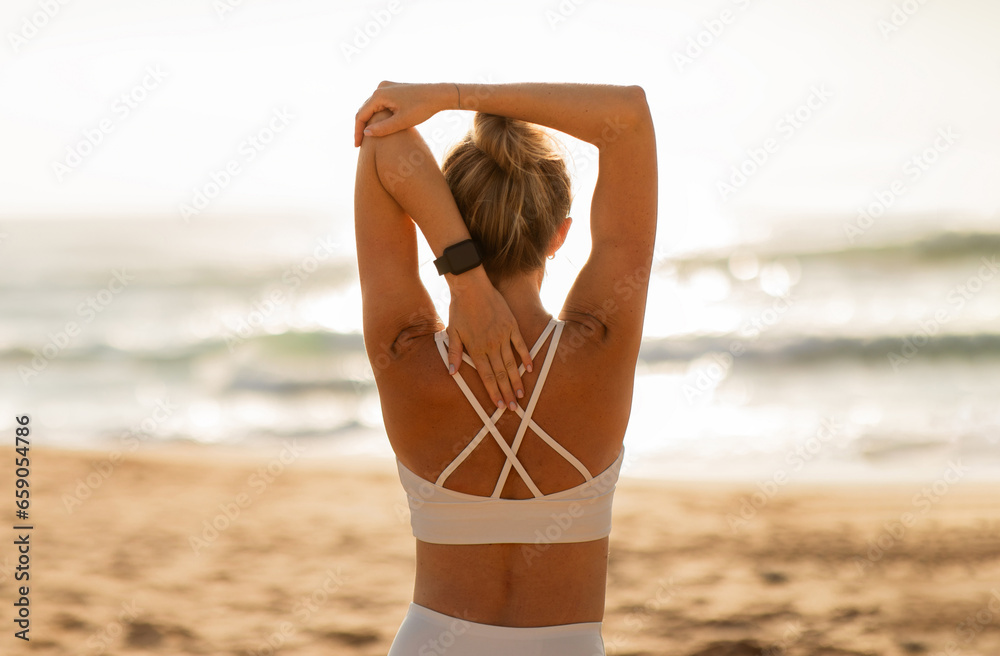 Back view of woman athlete in sportswear doing stretching and warm up for hands outdoors on ocean beach - obrazy, fototapety, plakaty 