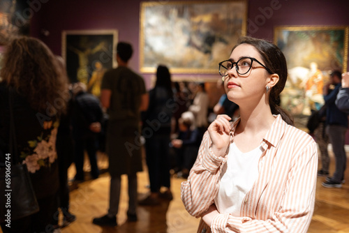 Portrait of thoughtful young Caucasian woman wearing glasses and looking at exhibition. In background, people are looking at paintings. Concept of Museum Day