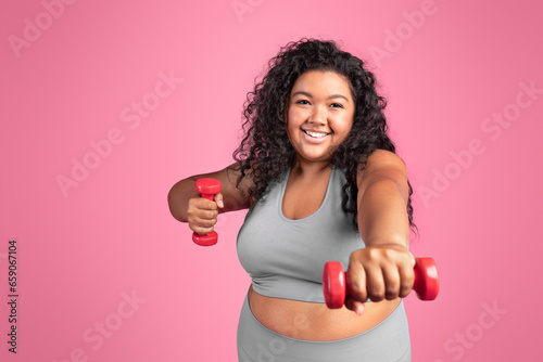 Cheerful excited black chubby lady in sportswear doing exercises with hand dumbbells, standing on pink studio background