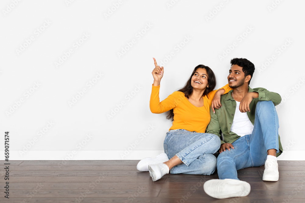 Happy young indian couple buying new house, sitting on floor in empty room, pointing at free space over white wall Happy young indian couple buying new house, sitting on floor in empty room, pointing at free space over white wall