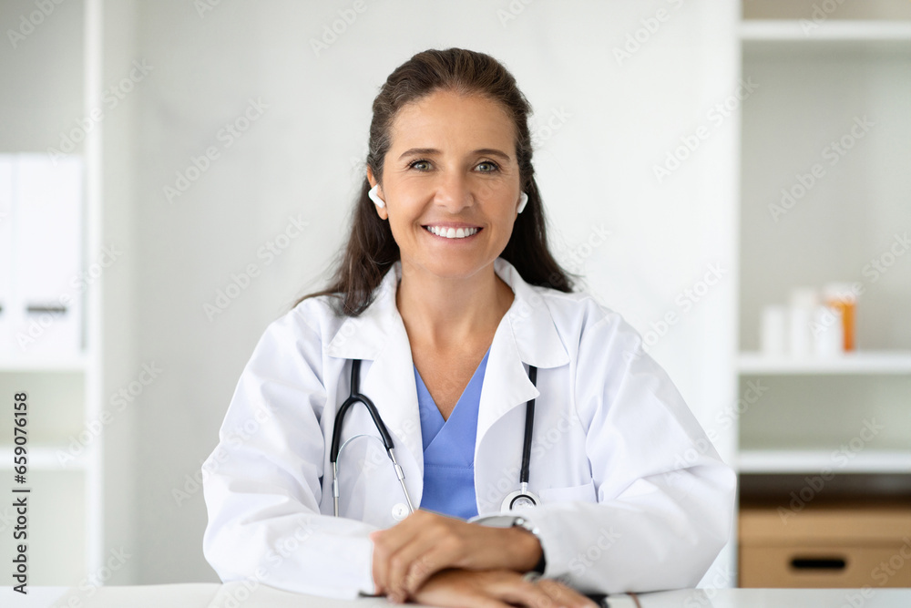 Positive mature lady doctor using wireless earphones. E-medicine, telehealth Positive mature lady doctor using wireless earphones. E-medicine, telehealth