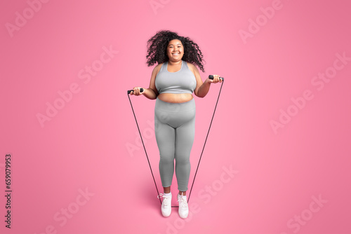 Happy african american chubby woman in sportswear jumping on jump rope, isolated on pink studio background, full length