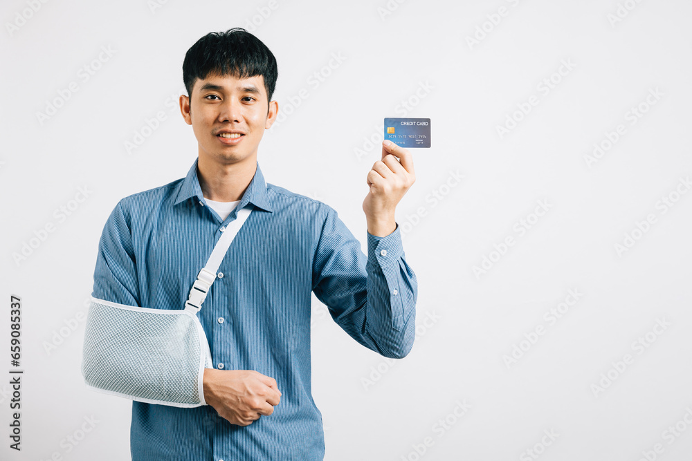 Despite a damaged arm, a man wears a support splint, paying medical bills with a credit card after an accident. Happy Asian man in a sling on white background, emphasizing health care. Copy space Despite a damaged arm, a man wears a support splint, paying medical bills with a credit card after an accident. Happy Asian man in a sling on white background, emphasizing health care. Copy space