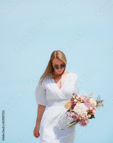 Joyful Young Woman Holding a Bouquet of Flowers on a Beach with Blue Sky Background