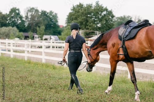 Female rider in equestrian clothes holding the reins and leading her beautiful saddled chestnut horse. Horseback riding activity concept.